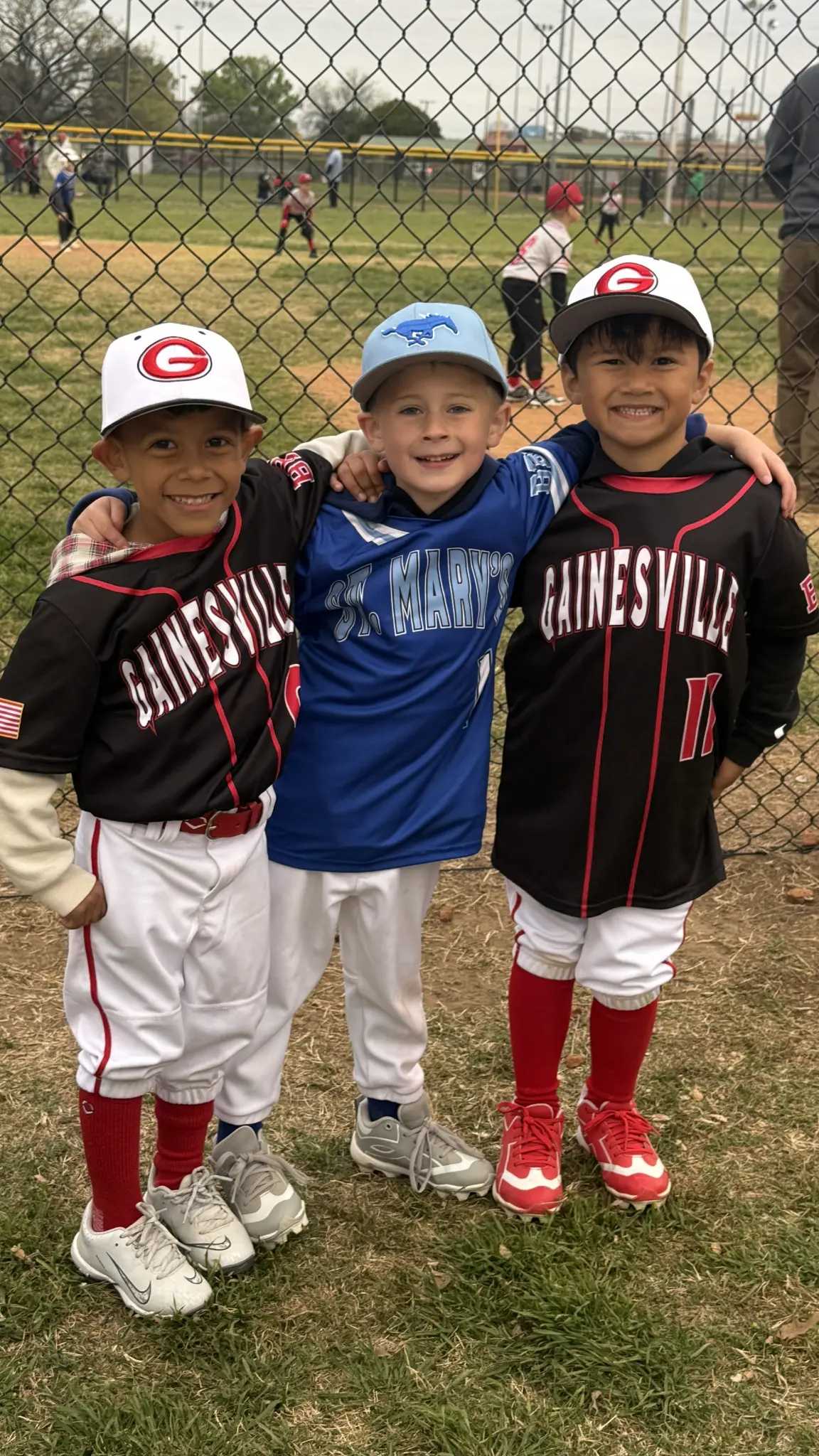 Three young baseball players smiling together beside the ballfield fence.